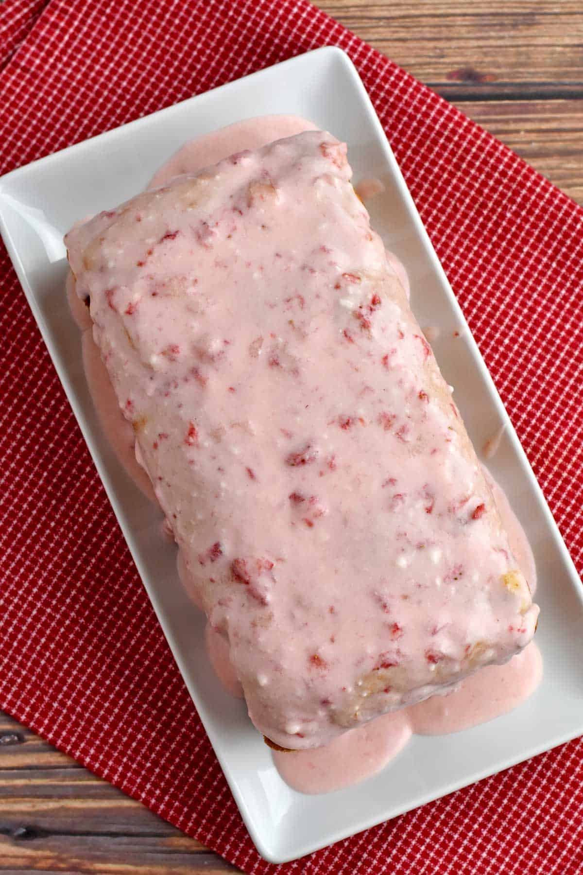 Glazed gluten free strawberry bread on white serving platter placed on red and white checkered kitchen towel.