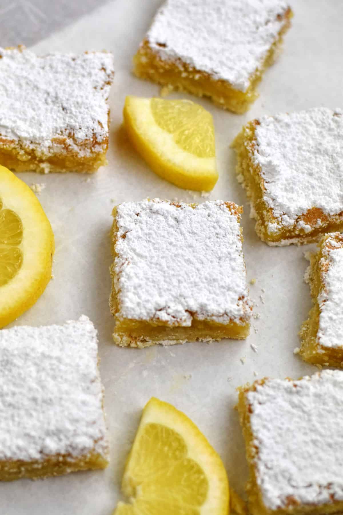 Gluten free lemon bars and lemon slices scattered on parchment paper.