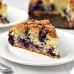 A slice of gluten free blueberry cake on a small white plate with more cake in the background.