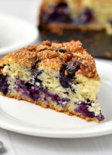 A slice of gluten free blueberry cake on a small white plate with more cake in the background.