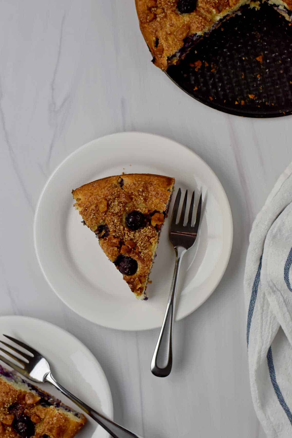 Overhead view of a slice of gluten free blueberry cake and a fork on a white dessert plate.