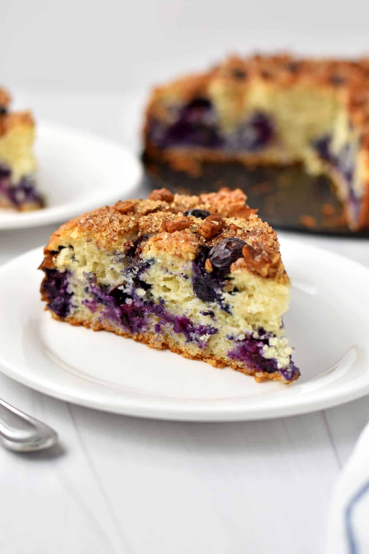 A slice of gluten free blueberry coffee cake on a small white plate with more cake in the background.